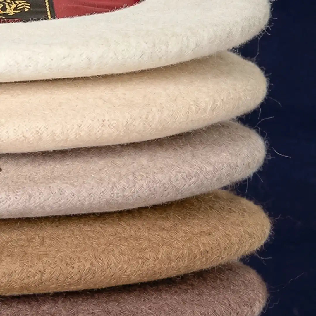 Gros plan sur les bords de bérets en laine empilés, montrant la texture et les coloris écru, beige, camel et taupe